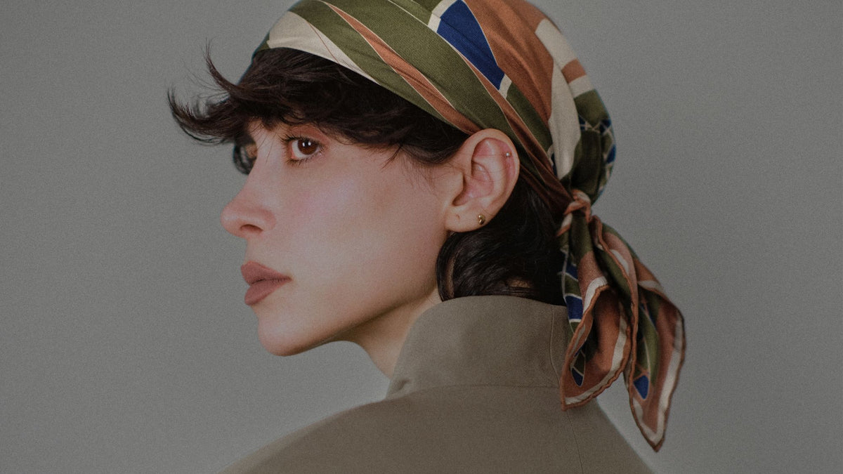 A woman in profile wearing a Tara Edition silk carré tied as a headscarf, over an olive jacket