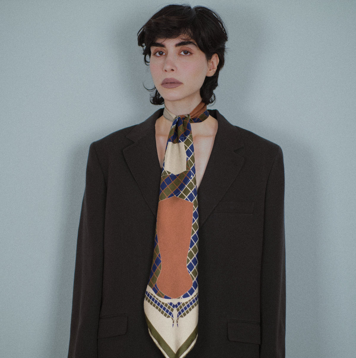 The Crossing silk carré worn folded long and knotted at the neck like a tie, over a dark jacket