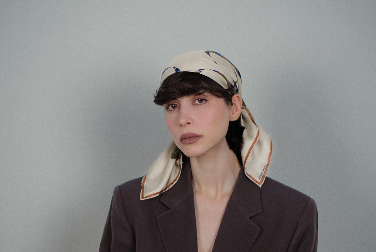 The Quatrefoil silk carré worn as a headscarf, the carob-bordered ends falling over the shoulders